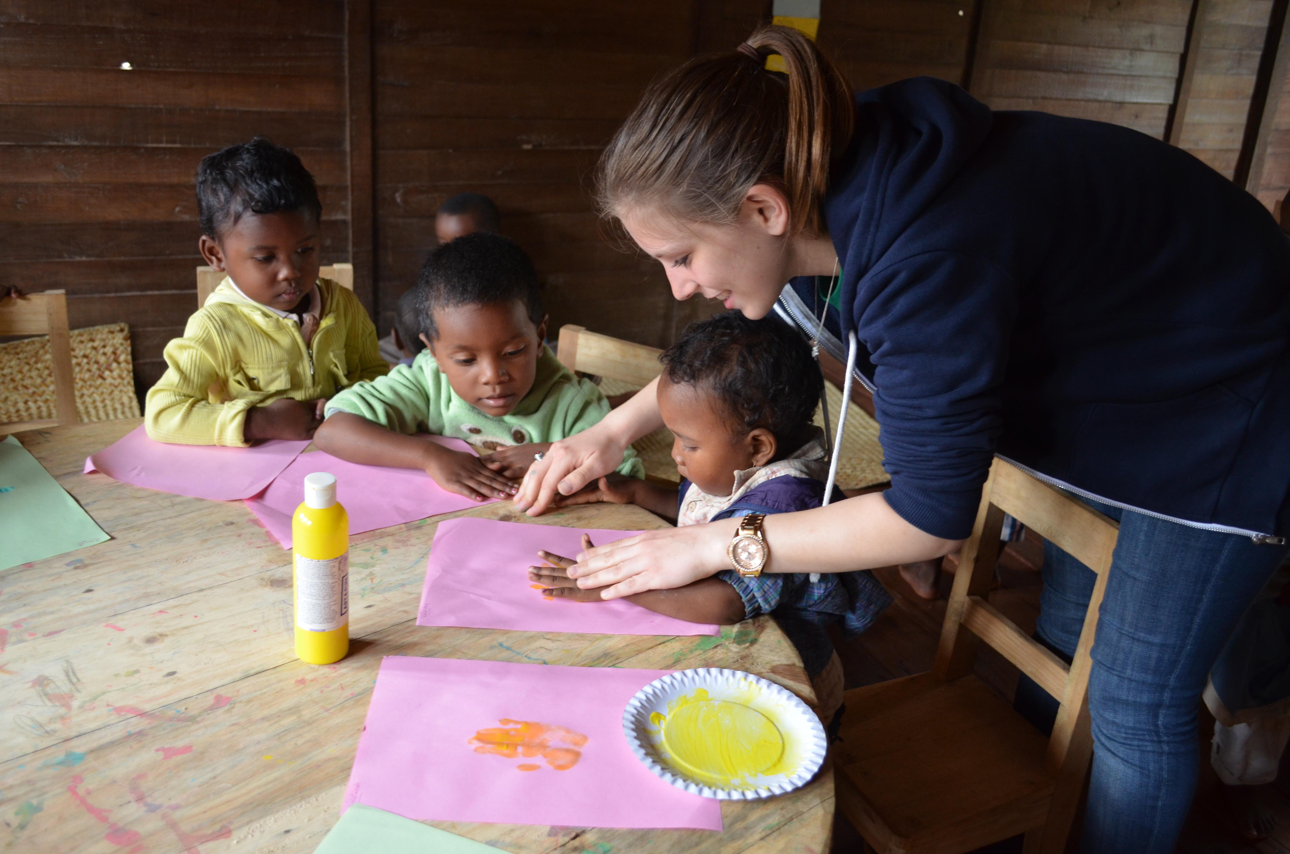 Projects Abroad Childcare volunteer helps a young child do finger painting during a creative activity at a care centre in Madagascar. Projects Abroad volunteer working with children in Madagascar helps a group of children do hand printing at a day care centre.